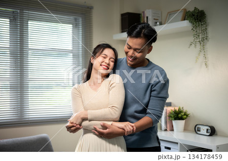 Sweet couple smiling asian man and woman holding hands while standing hugging in living room at home 134178459