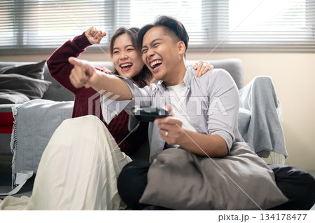Asian man holding console playing game and pointing aside a cheering woman on a living room's floor. Asian man holding console playing game and pointing aside a cheering woman on a living room's floor. 134178477