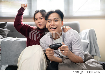 Asian man holding a console playing game with a woman hugging his shoulder on living room's floor. 134178478