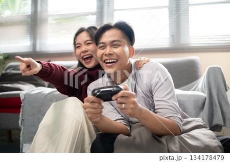 Asian man holding a console playing game with a woman pointing aside sitting on living room's floor. Asian man holding a console playing game with a woman pointing aside sitting on living room's floor. 134178479