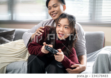 Asian woman holding a console playing game while lying over a man's lap on sofa in a living room. 134178483