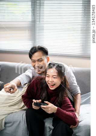 Asian man pointing and woman holding game console while lying over a man's lap on living room's sofa 134178485