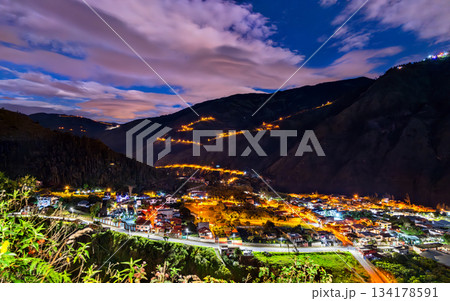Illuminated winding road of Via a Runtun rises above Banos de Agua Santa at night in Ecuador. Scenic landscape features glowing city lights and dark Andean mountains 134178591