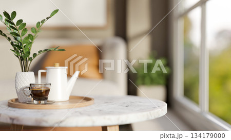 Teapot and glass cup with plant on wooden tray over round marble table across sofa in living room. 134179000