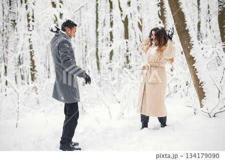 Indian man and caucasian woman walking in winter forest together. Lovely couple playing in snowballs. Man wearing grey coat and woman beige coat. Indian man and caucasian woman walking in winter forest together. Lovely couple playing in snowballs. Man wearing grey coat and woman beige coat. 134179090