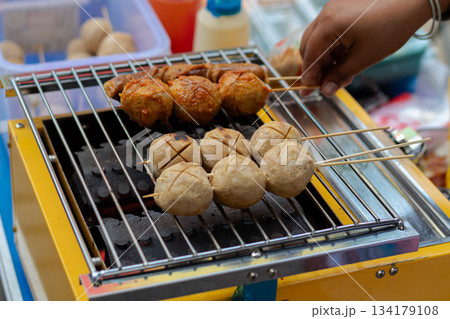 Close up of vendor grilling skewered street food meatballs and sausages on yellow charcoal barbecue. 134179108