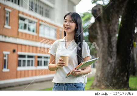 Young asian woman student holding book on laptop and coffee cup standing aside the building's garden Young asian woman student holding book on laptop and coffee cup standing aside the building's garden 134179365