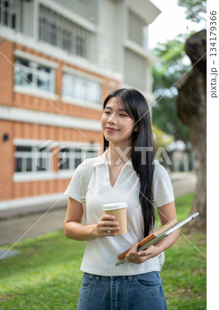 Young asian woman student holding book on laptop with coffee cup standing aside a building's garden. Young asian woman student holding book on laptop with coffee cup standing aside a building's garden. 134179366