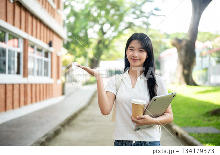 Young asian woman student holding laptop and coffee with hand open palm showing around the building. 134179373