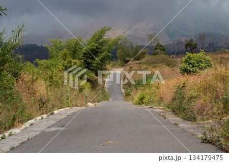 Empty asphalt road leading steeply uphill into misty mountains and thick dark clouds 134179475
