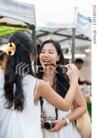 Asian woman feeding local food to her best friend who is standing holding a camera in local market. 134180142