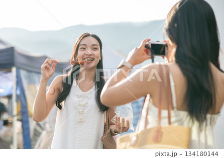 Best friend holding camera taking photo of asian woman standing and eating a food in local market. Best friend holding camera taking photo of asian woman standing and eating a food in local market. 134180144