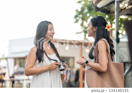 Pretty asian woman holding camera and talking with best friend while standing in the local market. Pretty asian woman holding camera and talking with best friend while standing in the local market. 134180145
