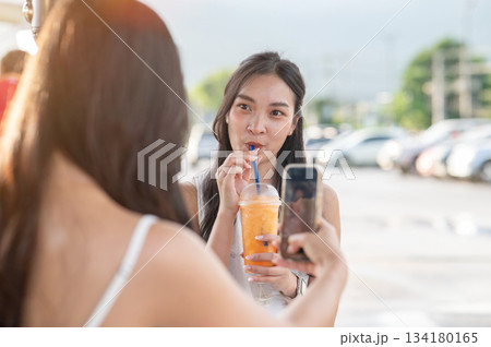 Pretty asian woman holding juice cup and looking at friend taking photo while sitting in local market 134180165