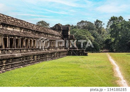 Wide angle view of massive stone wall and gallery of Angkor Wat temple with green grass field and footpath leading to forest 134180285
