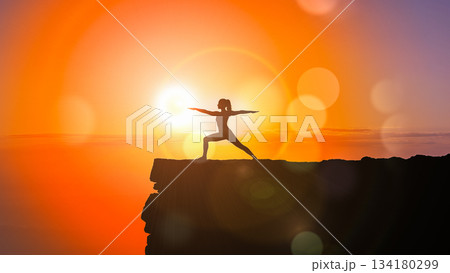 Silhouette of woman practicing yoga warrior pose on mountain cliff edge against orange sunset sky background illustrating freedom concept 134180299