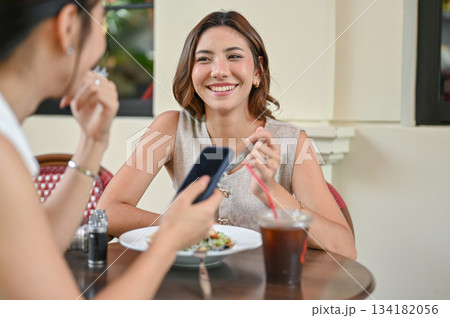 Asian woman holding fork talking with friend holding phone at wood table in front of restaurant cafe Asian woman holding fork talking with friend holding phone at wood table in front of restaurant cafe 134182056