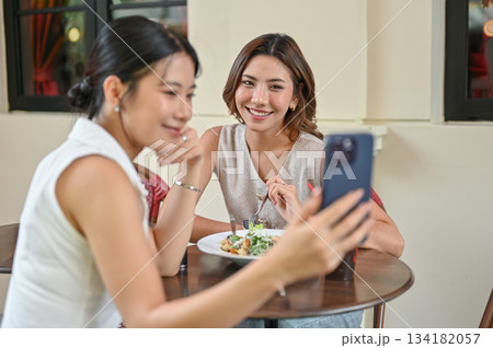 Asian woman holding phone taking picture with friend sit at wooden table in front of restaurant cafe 134182057