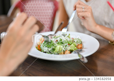 Close up woman's hands holding fork eating salad with friend sits at wooden table in restaurant cafe Close up woman's hands holding fork eating salad with friend sits at wooden table in restaurant cafe 134182058