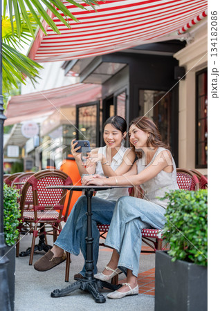 Pretty asian women taking picture or showing phone to friend while sitting at table outside the cafe 134182086