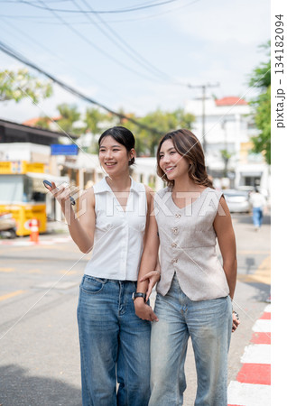 Pretty asian woman holding phone and pointing aside friend while standing or walking by street road. 134182094