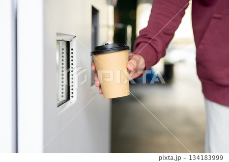 Close up of man's hand taking coffee or hot drink cup from automated beverage maker vending machine. 134183999