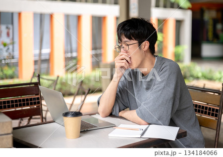 Glasses asian man sleepy yawning in front of laptop aside coffee and notebook while sitting at table 134184066