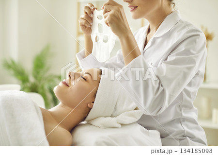 Female beautician applying moisturizing sheet mask on young woman face in beauty spa salon. 134185098