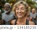 A joyful senior woman smiles brightly during an outdoor fitness activity, surrounded by friends jogging in the background, embodying health, energy, and vitality. 134185230