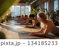 A serene yoga class practices on mats in warm sunrise light, with students aligned in a peaceful studio filled with plants and soft, calming atmosphere. 134185233