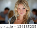 A smiling blonde student with bright blue eyes and a natural, fresh look stands confidently in a classroom setting, holding notebooks as soft light highlights her features. 134185235