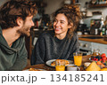 A young couple enjoys a cozy breakfast together, smiling warmly at each other in a relaxed kitchen setting filled with pastries, fruit, and fresh orange juice. 134185241