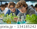 Two teenage students conduct a plant science experiment in a bright lab, observing seedlings and test tubes with curiosity while wearing protective goggles. 134185242