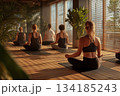 A group of people meditate peacefully in a sunlit yoga studio, sitting in calm stillness as warm light filters through the windows, creating a serene atmosphere. 134185243
