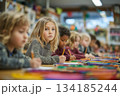 Children sit together in a colorful classroom, focused on drawing and writing. One child looks up thoughtfully, capturing a moment of learning and creativity. 134185244