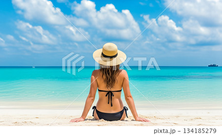 Rear view woman sitting white sand beach wearing straw hat black bikini looking turquoise ocean water summer vacation travel holiday tropical relax 134185279