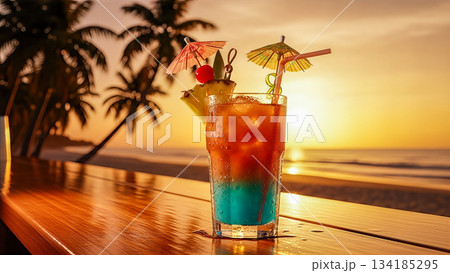 Tropical cocktail glass with umbrella and fruit on wooden bar counter against beautiful ocean sunset background during summer vacation 134185295