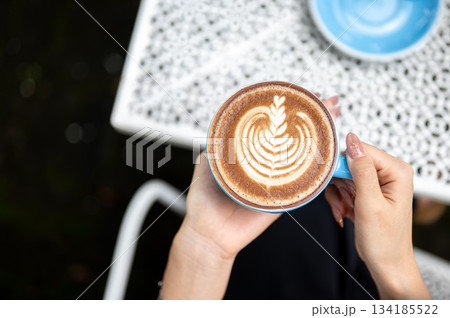 Close up a woman's hands holding latte art coffee in blue ceramic cup over floral pattern cafe table 134185522