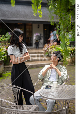 Pretty asian woman standing with crossing arms looking at a glasses man sits at table in cafe garden Pretty asian woman standing with crossing arms looking at a glasses man sits at table in cafe garden 134185538