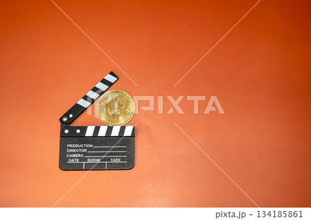 A Bitcoin coin rests on a film clapperboard against an orange background. This image suggests cryptocurrencies' potential in film production. 134185861