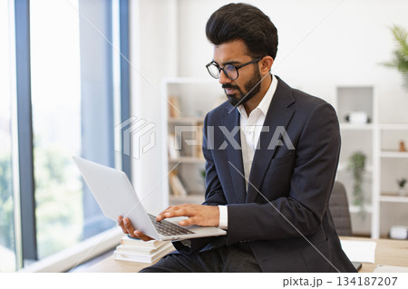 An Indian businessman wearing glasses works on his laptop in a modern office setting An Indian businessman wearing glasses works on his laptop in a modern office setting 134187207
