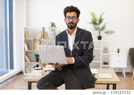 An Indian businessman in a suit is working on his laptop while sitting on a desk in an office setting 134187232