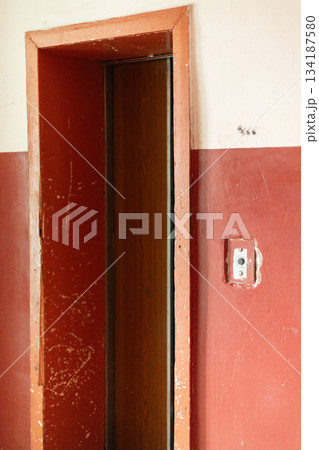 Inside an old house, a weathered doorway stands alone. The walls are peeling with faded red paint, revealing years of disrepair and neglect 134187580