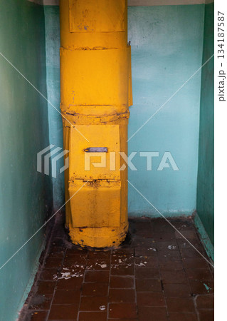 Inside an old building, a yellow garbage chute stands against a wall. The surrounding walls are in a faded blue, and the floor shows signs of wear and dirt 134187587