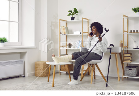 Young woman with injured leg sitting at home on chair with crutches and talking on the phone. 134187606