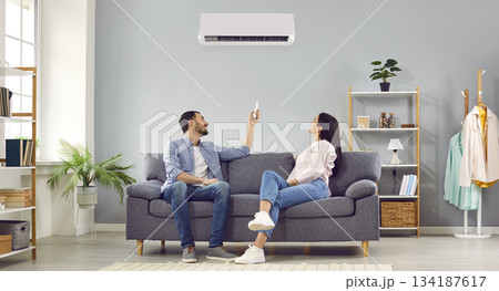 Young happy couple turning on air conditioner sitting on sofa at home. Banner 134187617