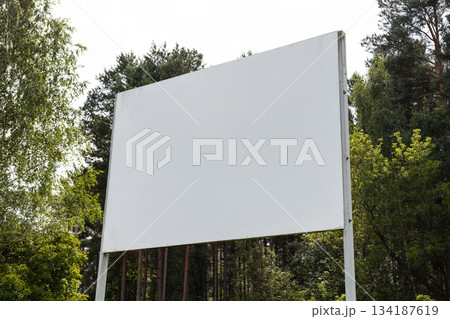 A large blank billboard is positioned in a green forest area surrounded by trees. The bright daylight illuminates the space, creating a peaceful atmosphere 134187619