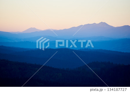 Serene view of rolling mountains in shades of blue and green, with distant peaks under soft, pastel sky, creating peaceful and tranquil landscape. 134187727