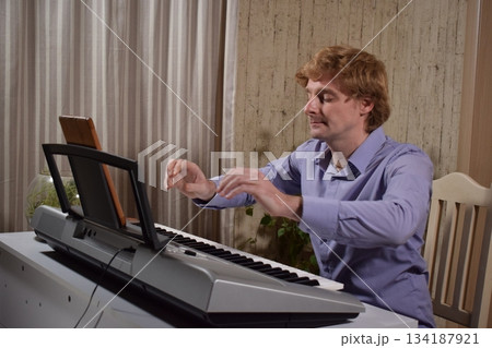 A young white man plays an electronic synthesizer indoors, immersed in his creative hobby. Focused on shaping tones and melodies, he explores musical expression and artistry in a calm home setting. 134187921