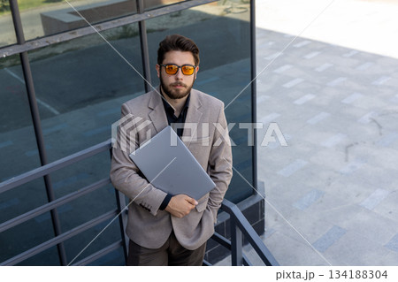 Stylish businessman works on laptop while standing outdoors, full length 134188304